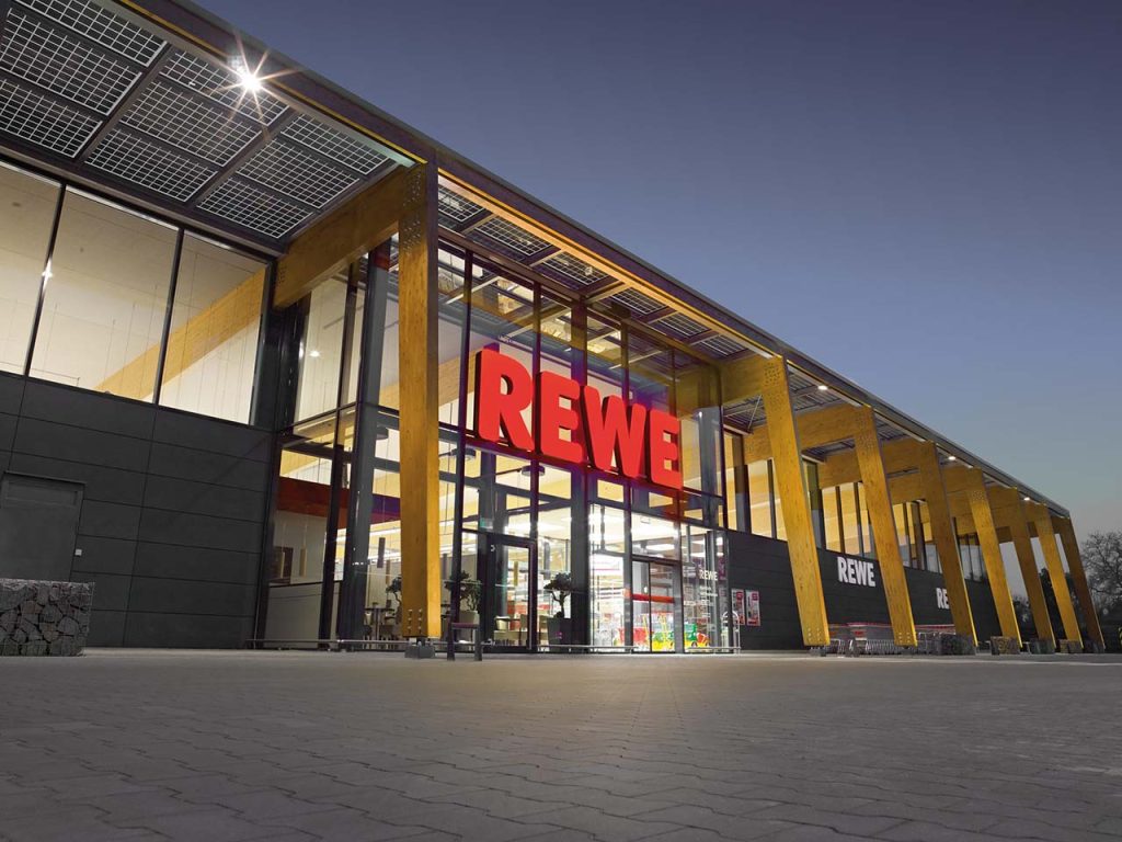 Eingangsbereich eines großen REWE-Store von außen mit dem roten REWE-Logo über dem Eingang