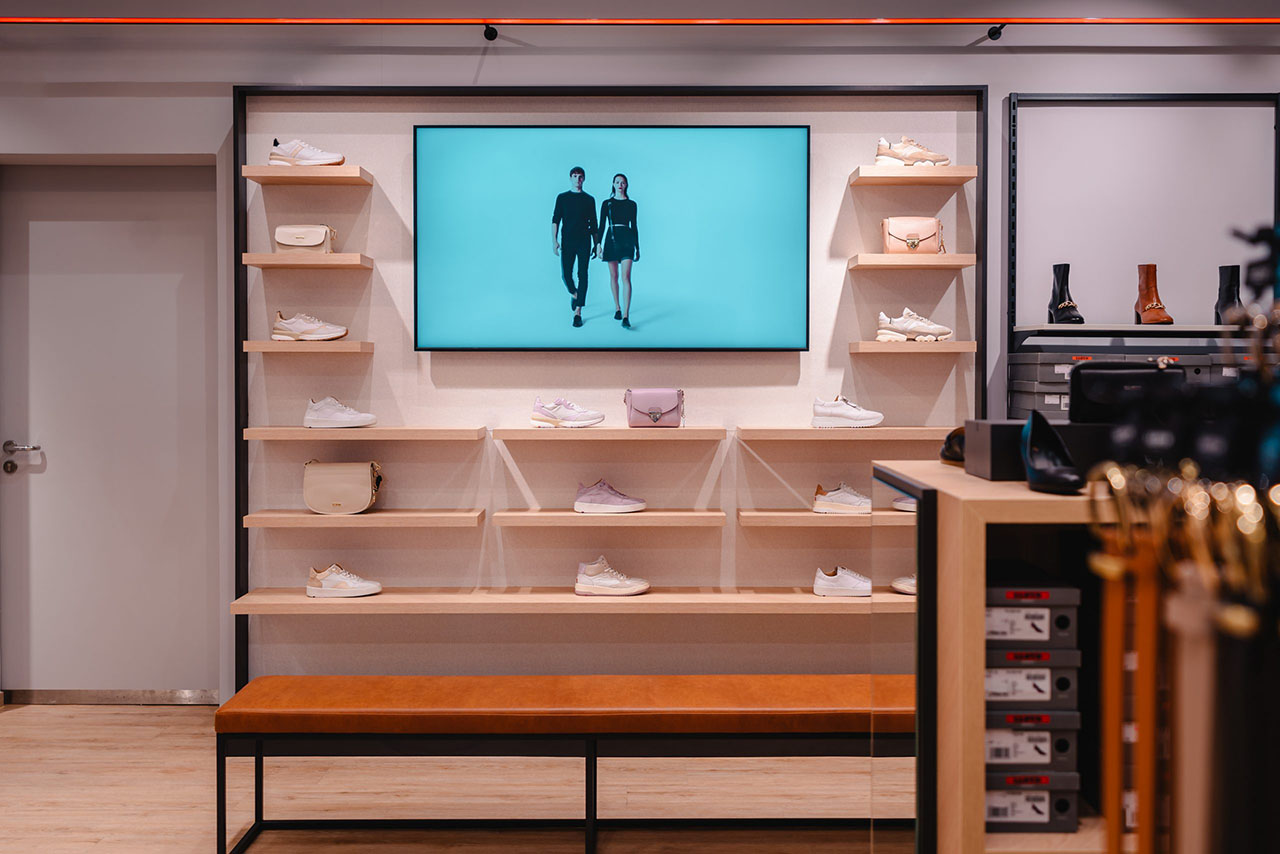 Sneakers, die auf Holzregalen an einer Wand im LLOYD-Store ausgestellt sind, mittig ein Screen im Querformat mit Content eines Paars