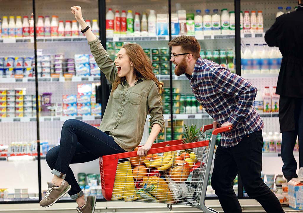 Ein Mann schiebt einen Einkaufswagen gefüllt mit Einkäufen, darauf sitzt eine Frau, beide haben Spaß