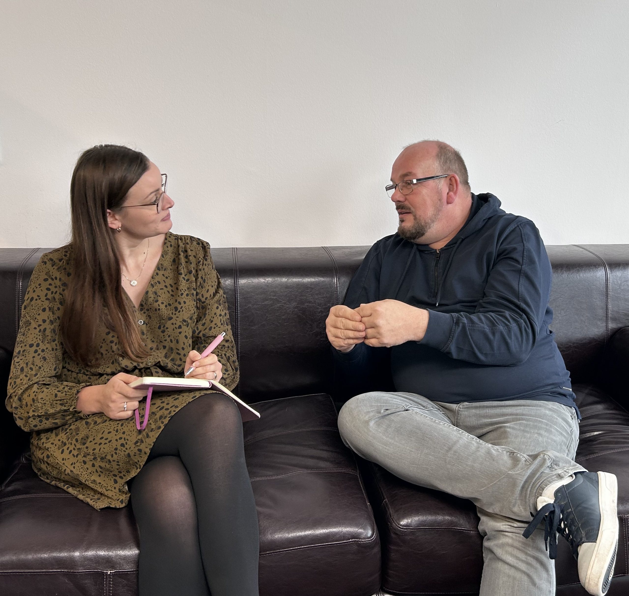 Zwei Personen sitzen auf der Couch und reden, eine Person mit Notizbuch in der Hand